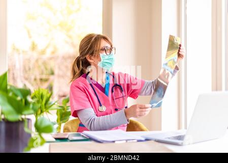 Aufnahme einer Ärztin, die Gesichtsmaske trägt, während sie im Zimmer des Arztes sitzt und das Röntgenbild überprüft. Stockfoto