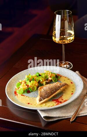 Schmackhaftes gegrilltes Seebarsch-Filet mit gedünsteten rosenkohl, gebratenen Pilzen, Kartoffeln, karamellisierten Zwiebeln und cremiger Sauce, serviert mit einem Glas Stockfoto