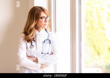 Porträtaufnahme einer denkenden Ärztin, die mit gekreuzten Armen am Fenster steht und aus dem Fenster schaut. Stockfoto