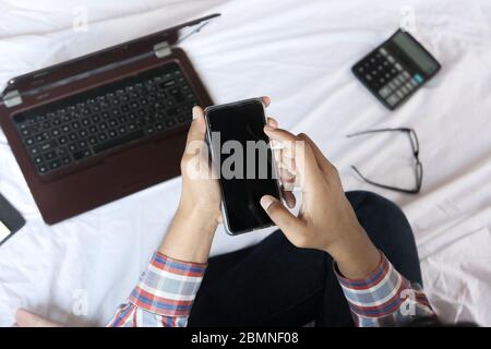 Junger Mann, der von zu Hause aus mit Smartphone auf dem Bett arbeitet Stockfoto