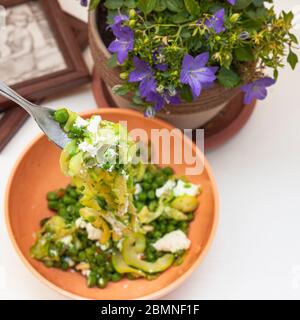 Zucchini Spaghetti mit Erbsen und Ricotta in natürlichem Licht mit einer schönen Pflanze mit lila Blüten und zwei kleine Plätze in der Nähe Stockfoto