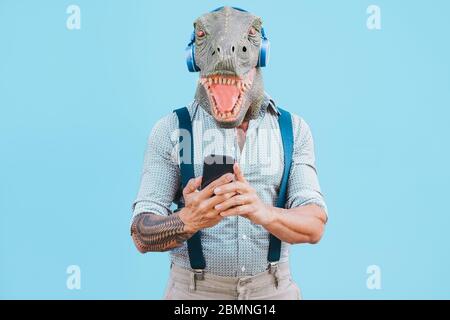 Senior tätowierten Mann mit t-rex Maske mit Smartphone während Musik während Coronavirus Zeit hören - Verrückter Mann trägt lustige Maske für Covid-19 verhindern Stockfoto