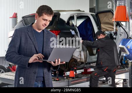 Manager Laptop halten, während in der Nähe Auto in der Werkstatt mit Mechaniker Reparatur Auto auf Hintergrund stehen Stockfoto