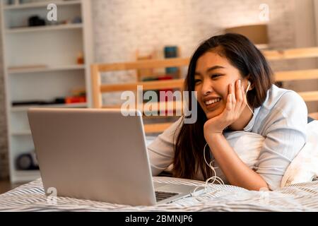 Junge lächelnde asiatische Frau in Freizeitkleidung und auf dem Bett mit Laptop-Computer liegen. Freiberufliche Mitarbeiterin, die von zu Hause aus am Notebook arbeitet Stockfoto