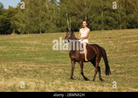 Junges Mädchen in weißem Kleid reiten auf Pferd ohne Sattel durch Wiese am Sommernachmittag Stockfoto