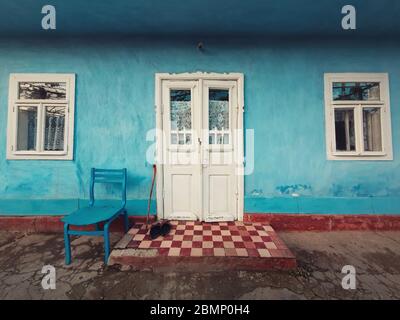 Stock und ein Paar alte Schuhe und ein Stuhl vor der Haustür eines alten Hauses. Traditionelle ländliche Gebäudefassade. Blau-lime bemalte Wände, weiß Stockfoto