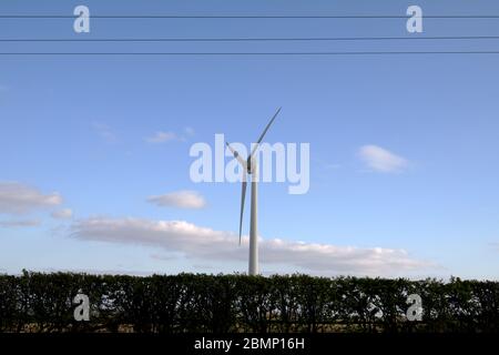 Eine dreiblättrige Windturbine in einem Feld außerhalb von Nottingham, England hinter einer Hecke mit blauem Himmel und fleckigen Wolken Stockfoto