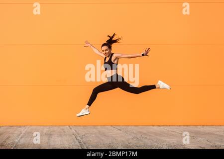 Positives lebhaftes Mädchen mit passender Körperform in straffer Sportbekleidung springen, laufen und lächeln voller Energie, gesunder Sportlebensstil. Outdoor-Studio-Schuss Stockfoto