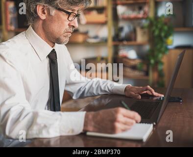 Geschäftsmann Schreiben auf einem Notebook, Mann mittleren Alters, der von zu Hause aus intelligent arbeitet, nutzt moderne Technologie, um auf Laptop und Smartphone zu kommunizieren, blurre Stockfoto