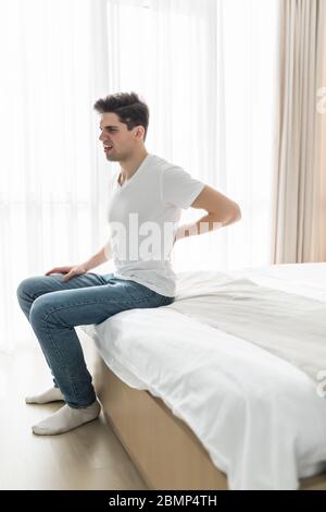 Porträt Des Jungen Mannes, Der Auf Dem Bett Sitzt Und An Rückenschmerzen Leidet Stockfoto