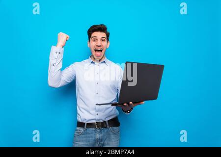 Emotional junger Mann mit Laptop feiert Sieg auf farbigen Hintergrund Stockfoto