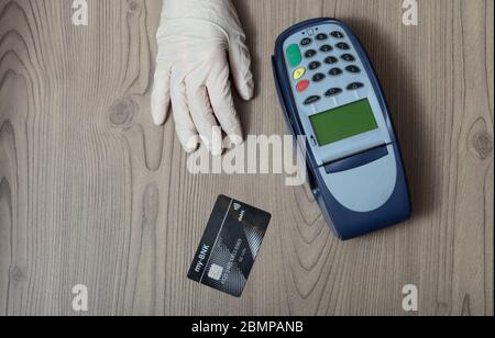 Hand in einem schützenden Einweg-Handschuh mit Bankkarte und Bankautomat auf einem hölzernen Ladentisch konzeptionell des Einkaufs während der Covid-19 Pandemie Stockfoto