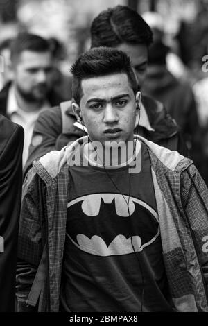 Junger Mann mit Batman-Shirt und kabelgebundenen Ohrhörern in der Menge, der direkt auf die Kamera in London, England, Großbritannien schaut Stockfoto