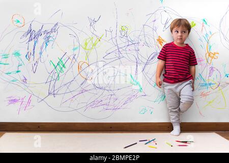 Unfug des Kindes. Junge mit einem abgelenkten Gesicht, weil er die ganze Wand gezeichnet. Kleiner Junge lehnte sich an die weiße Wand, wo er viele Zeichnungen machte Stockfoto