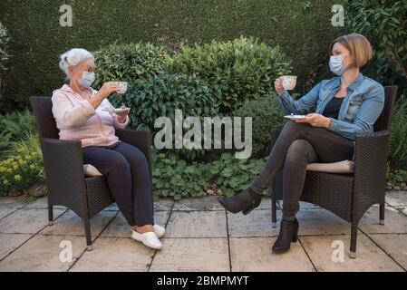 Ältere Frau und Tochter mit Gesichtsmasken mit Kaffee im Garten auf Safetyl Entfernung Stockfoto