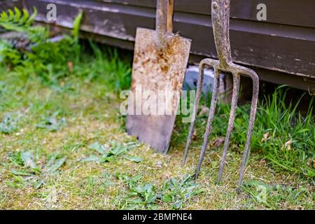 33 Nahaufnahme von rostigen Gartengabeln und Spaten, die sich gegen einen Holzschuppen in einem englischen Landgarten mit absichtlich geringer Schärfentiefe lehnen Stockfoto