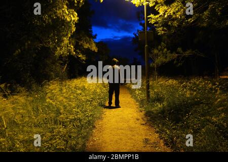 Eine geheimnisvolle Kapuzenfigur, die nachts mit dem Rücken zur Kamera unter einer Straßenbeleuchtung steht Stockfoto