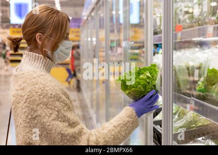 Frau Käufer trägt medizinische Maske und Latexhandschuhe, wählt Salat Salatblätter durch Öffnen Kühlschrank im Supermarkt. Schutzmaßnahmen gegen Stockfoto