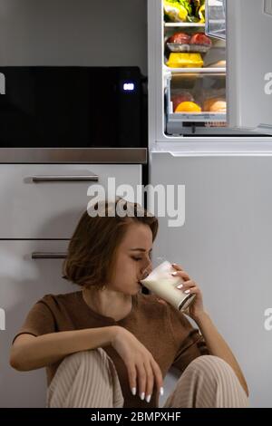 Junge Frau, die um Mitternacht Milch in der Nähe des offenen Kühlschranks trinkt, während sie in der Küche auf dem Boden sitzt. Night Eating Syndrom Stockfoto