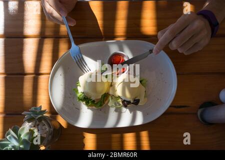 Person, die Eier benedikt mit Hollandaise Sauce und Lachs mit halbierten Kirschtomaten serviert. Frühstück im Freien. Draufsicht Stockfoto