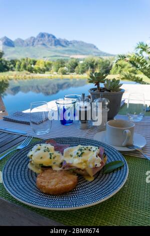 Eier Benedikt mit Hollandaise und Rösti serviert auf einem Teller draußen. Frühstück mit pochierten Eiern im Freien und Blick auf die Berge Stockfoto