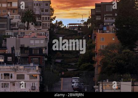 Die berühmte krumme Lombard Street in San Francisco, Kalifornien, mit einer kultigen Seilbahn auf dem Hügel und einem feurigen Himmel bei Sonnenuntergang Stockfoto