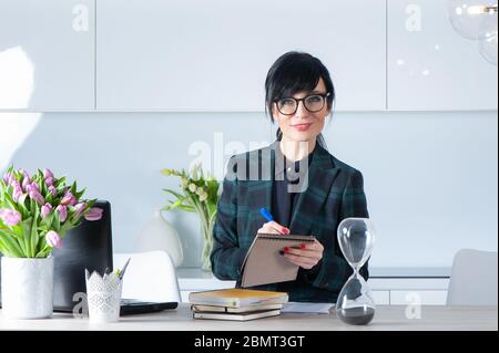 Schreiben Sie es auf. Schönes Mädchen in Anzug und Brille an ihrem Schreibtisch, hält einen Stift und ein Notizbuch, schaut auf die Kamera und lächelt. Zufrieden mit t Stockfoto