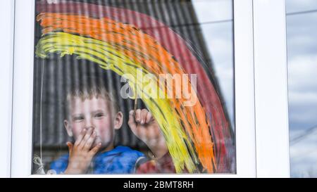 Kleiner Junge, der Regenbogen an einem Fenster malt und durch das Fenster schaut, während er während der Quarantäne zu Hause sitzt. Stockfoto