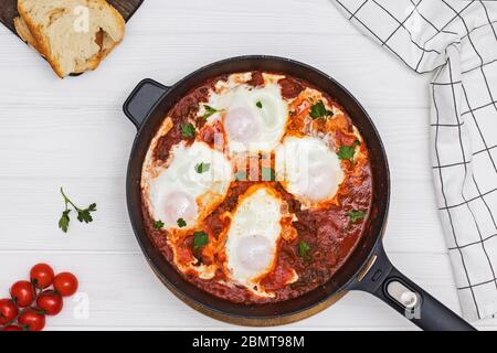 Shakshuka Eiergericht mit Tomatensauce in einer schwarzen Gusseisenpfanne auf dem weißen Holztisch Stockfoto