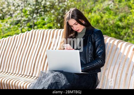 Junge elegante Frau auf Bank im Park sitzen und arbeiten auf Laptop an einem sonnigen und windigen Tag, das Konzept der Arbeit außerhalb des Büros Stockfoto