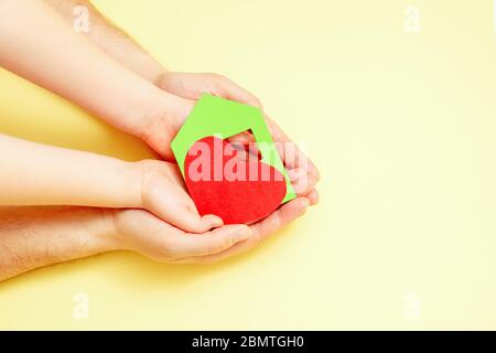 Papier grünes Haus mit rotem Herz in Papa und Kind die Hände auf gelbem Hintergrund. Wohltätigkeitskonzept. Stockfoto