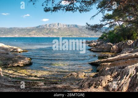 Kleiner versteckter Strand in einer Bucht auf dem Hügel Marjan in Split, Kroatien. Schönes Meer im Schatten, Berge und Küstenstädte in der Ferne gesehen Stockfoto