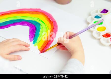 Nahaufnahme der Hand des Kindes mit bemalten Regenbogen auf weißem Papier während der Covid-19 Quarantäne zu Hause. Kreativität für Kinder. Stockfoto