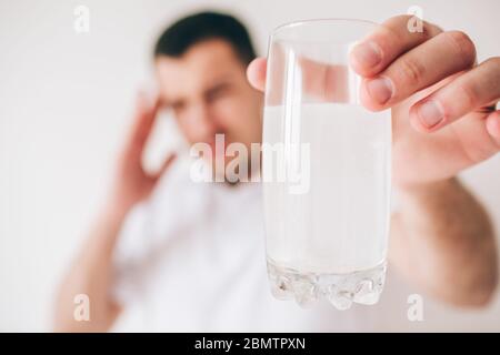 Junger kranker Mann isoliert über Hintergrund. Guy halten Glas Wasser mit Brausetabletten und Medizin darin. Leiden Sie unter Kopfschmerzen und Schmerzen. Verschwommene BA Stockfoto