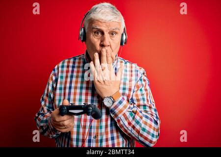 Senior gut aussehend hoary Gamer Mann spielen Videospiel mit Joystick und Kopfhörer decken Mund mit Hand schockiert mit Scham für Fehler, Ausdruck f Stockfoto