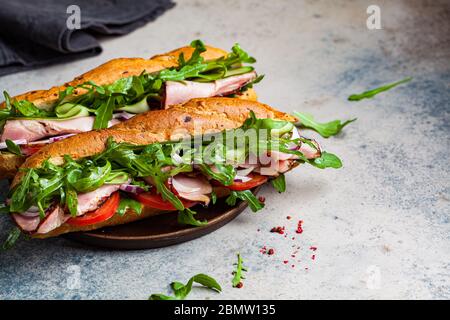 Zwei frische Baguette-Sandwiches mit Fleisch, Tomaten, Gurken und Rucola auf grauem Hintergrund. Stockfoto