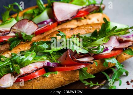 Zwei frische Baguette-Sandwiches mit Fleisch, Tomaten, Gurken und Rucola auf grauem Hintergrund. Stockfoto