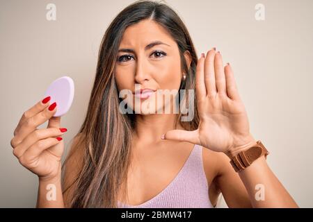 Junge schöne Frau mit Schönheit Schwamm mit Gesichtsmaske über rosa Hintergrund mit offener Hand tun Stop-Zeichen mit ernsten und selbstbewussten Ausdruck Stockfoto