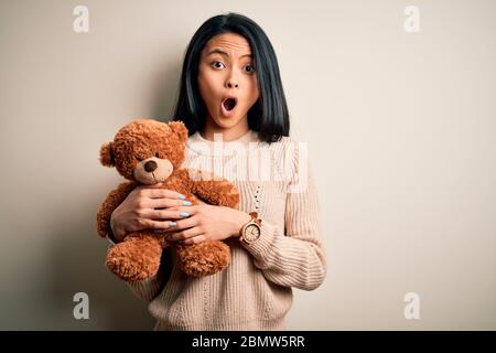 Junge schöne chinesische Frau mit Teddybär stehend über isoliert weißer Hintergrund Angst im Schlag mit einer Überraschung konfrontiert, ängstlich und aufgeregt mit Stockfoto