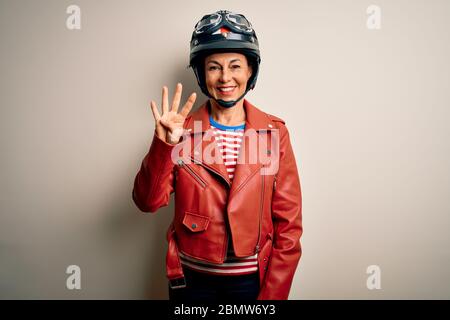 Motorradfahrerin im mittleren Alter, die Motorradhelm und Jacke auf weißem Hintergrund trägt und mit den Fingern Nummer vier zeigt, während Smilie Stockfoto