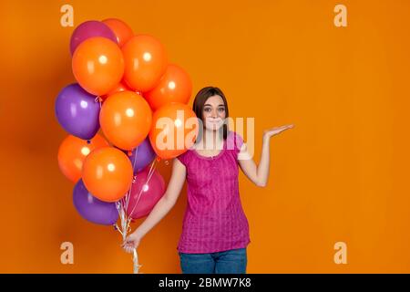 Zweifelhaft verwirrt Frau in rosa T-Shirt posiert mit hellen bunten Luftballons isoliert auf orangefarbenem Hintergrund. Geburtstagsfeier. Copy space Stockfoto