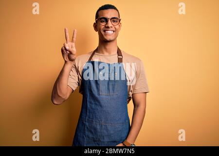 Junge gut aussehende afroamerikanischen Ladenbesitzer Mann trägt Schürze über gelbem Hintergrund lächelnd mit glücklichem Gesicht zwinkerte an der Kamera tun Siegeszeichen. Stockfoto