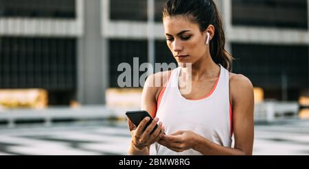 Frau in Sportbekleidung mit ihrem Smartphone nach dem Training Sitzung. Gesunde Frau mit Ohrhörern, die eine Pause vom Training einlegen und ihre Fitness überprüfen Stockfoto