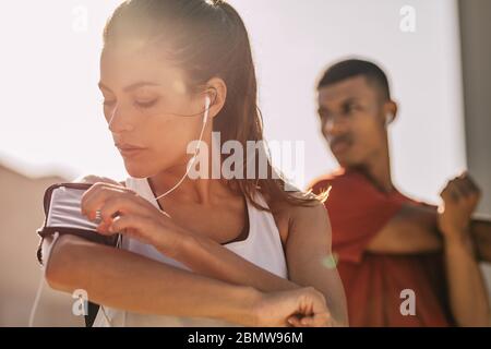 Nahaufnahme einer Frau, die mit ihrem Smartphone Musik hört, während sie in der Stadt mit einem Mann im Hintergrund trainiert. Frau mit einem Smartp Stockfoto