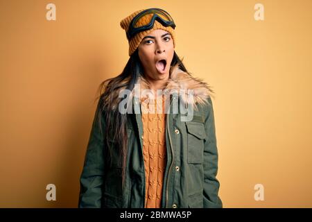 Junge schöne hispanische Frau mit Skibrille und Mantel für Winterwetter in Schock Gesicht, sah skeptisch und sarkastisch, überrascht mit offenen mou Stockfoto