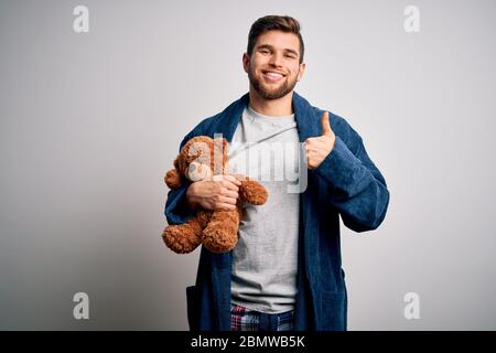 Junge blonde Mann mit Bart und blauen Augen tragen Pyjama halten Teddybär glücklich mit großen Lächeln tun ok Zeichen, Daumen mit Fingern, ausgezeichnetes Zeichen Stockfoto