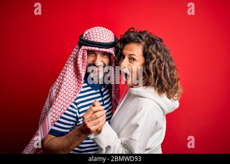Schönes Paar im mittleren Alter mit arabischem Kopftuch und Sweatshirt, lächelnd glücklich und selbstbewusst. Mit einem Lächeln auf dem Gesicht stehend, umarmend über isoliertem Rot B. Stockfoto