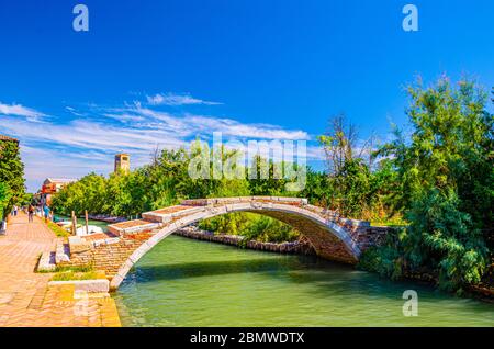 Steinteufel Brücke über den Wasserkanal auf der Insel Torcello, Uferpromenade entlang des Wasserkanals, grüne Bäume, Turm und blauer Himmel Hintergrund. Lagune Von Venedig, Region Venetien, Norditalien. Stockfoto