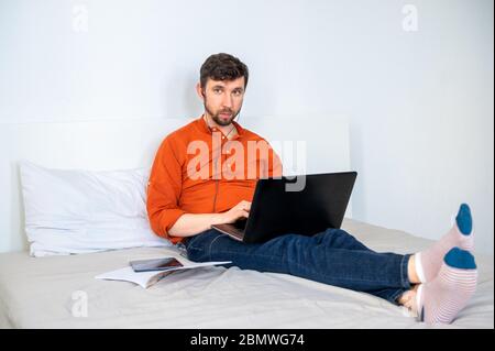 Ein ernsthafter Mann, der zu Hause mit einem Laptop und einem Headset auf dem Bett arbeitet Stockfoto