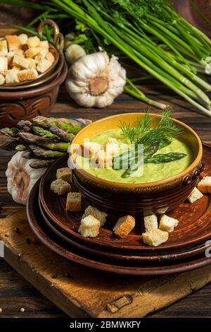 Cremige Spargelsuppe in Glasschale auf schwarzem Teller vor rohem frischem Spargel und Grün auf rustikalem Holzhintergrund Stockfoto